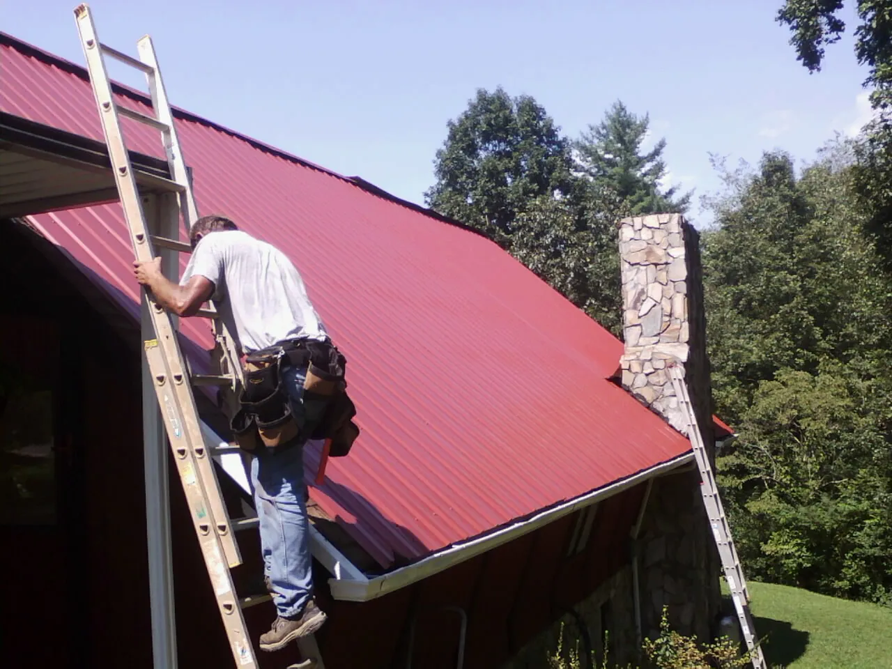 Roofer on a ladder beside a red metal roof and stone chimney — Soffit & Fascia Repair by Roofing Brothers in Yanceyville